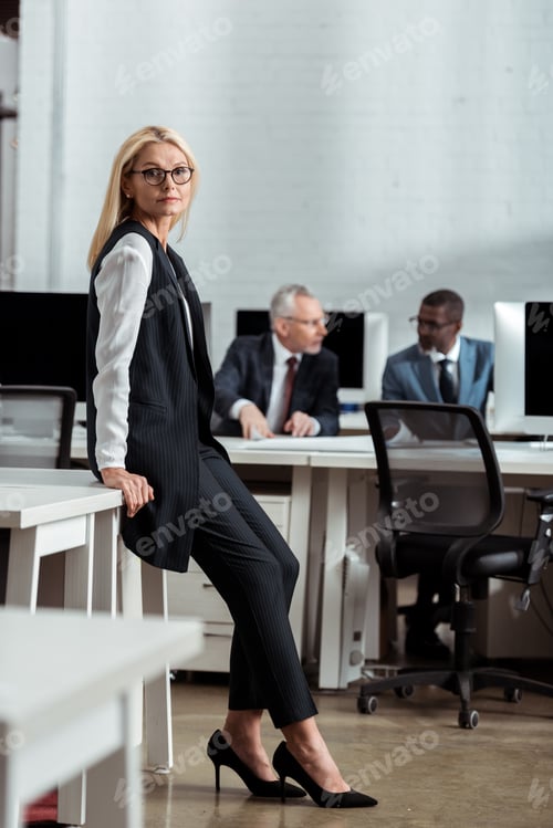 Preview: selective focus of attractive businesswoman in glasses looking at camera near multicultural men