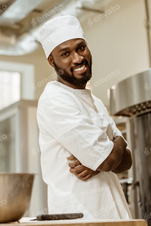 Preview: handsome african american baker with crossed hands looking at camera on baking manufacture