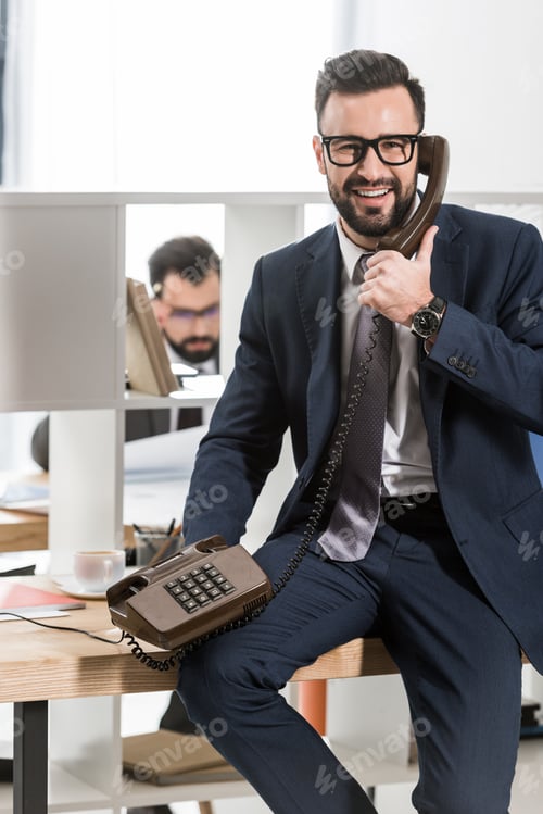Preview: smiling businessman talking by stationary telephone in office and looking at camera