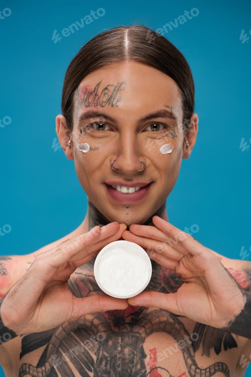Preview: happy young man with tattoos and cream on cheeks holding container isolated on blue