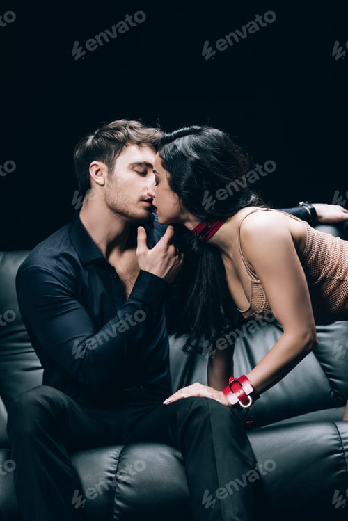 Preview: sexy brunette woman in beige underwear and red handcuffs kissing man on leather sofa isolated on