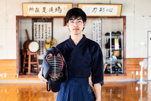 Preview: Male Japanese Kendo fighter standing in a gym, holding Kendo mask, looking at camera.