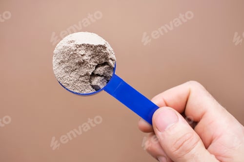Preview: Male hand holds a measuring spoon with brown protein on brown background top view.