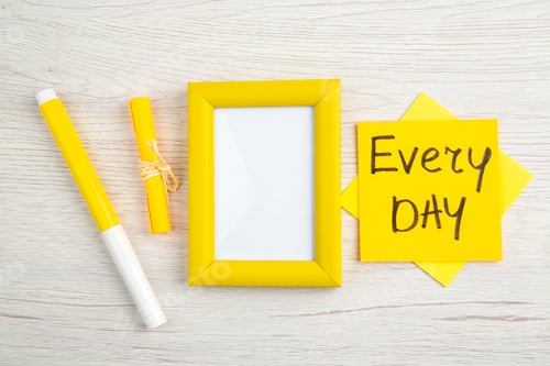 Preview: Overhead view of yellow empty picture frame and every day note markers on white stripped background