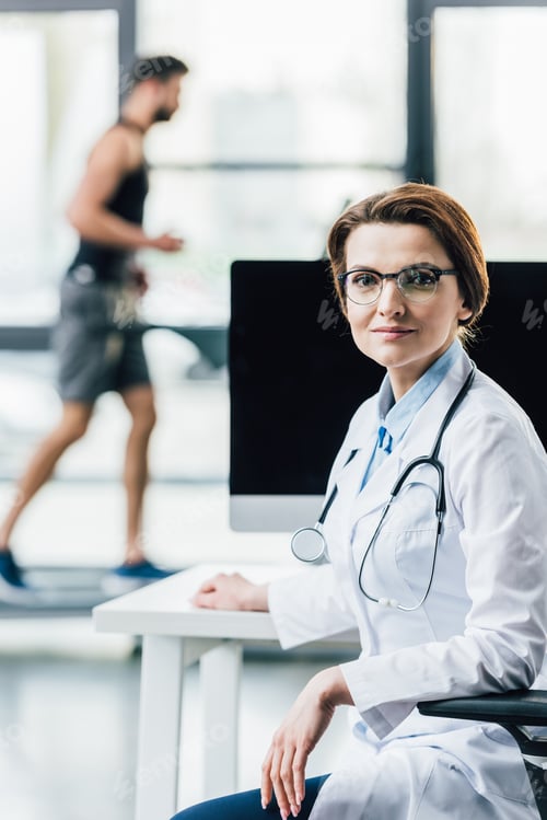 Preview: beautiful doctor sitting at computer desk in gym and looking at camera