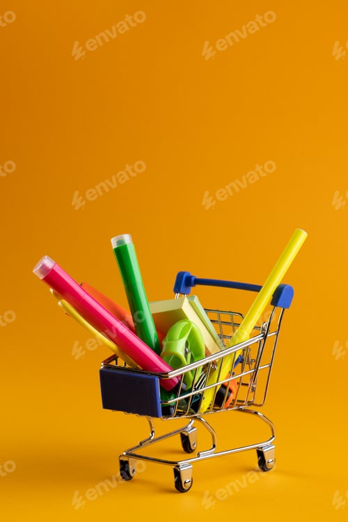 Preview: Close up of miniature shopping trolley with school materials and copy space on orange background
