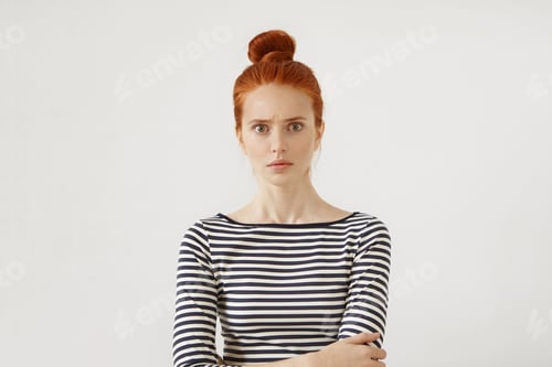 Preview: Serious female model with reddish hair tied in bun, standing against white background in casual swea