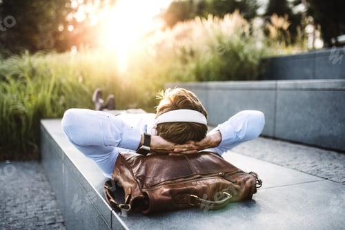Preview: A businessman with headphones, lying on the steps outdoors at sunset.