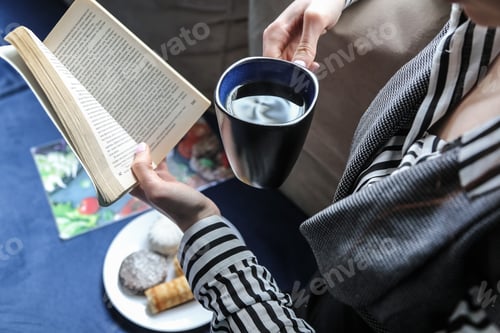 Preview: side view woman drinking cup of tea with cookies and reading a book