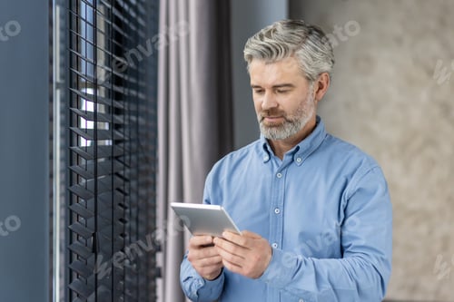 Preview: Senior gray-haired serious male businessman stands in the office by the window and uses a tablet