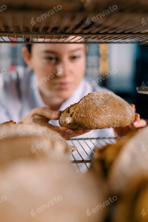 Preview: attractive female baker between shelves looking checking freshly baked bread very carefully