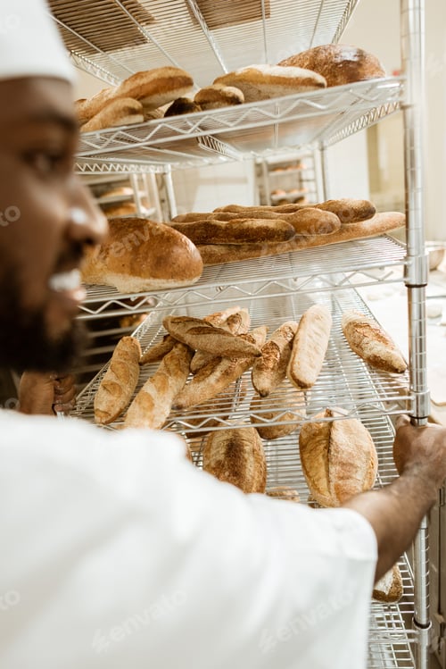 Preview: close-up shot of african american baker driving shelves of fresh bread on baking manufacture