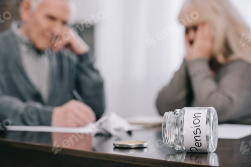 Preview: selective focus of 'pension' word on empty glass jar with senior couple sitting at table on
