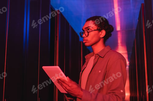 Preview: young african american programmer with digital tablet working near servers in data center in neon