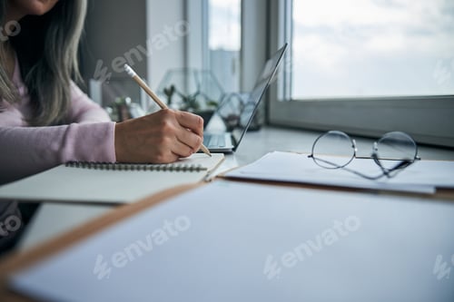 Preview: Woman writing on notebook and using laptop at home