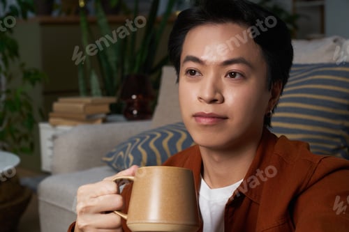 Preview: Asian man in a cozy modern apartment with a warm drink in hand
