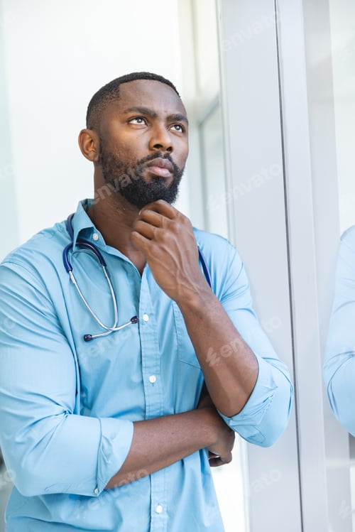 Preview: Thoughtful african american male doctor wearing blue shirt looking through window in hospital room