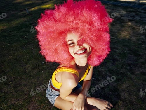 Preview: Redhaired woman sitting on the ground, hands in her hair, smiling broadly, outdoors, casual outfit