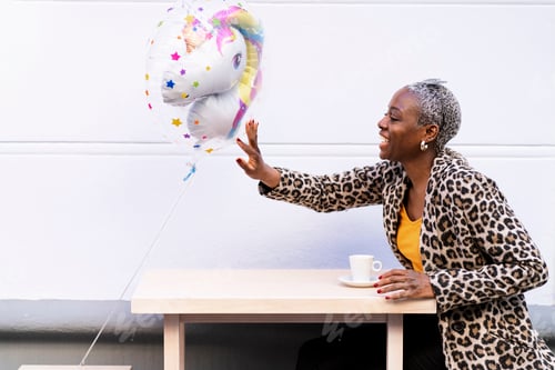 Preview: Side view of positive African American female touching colorful unicorn balloon while sitting at