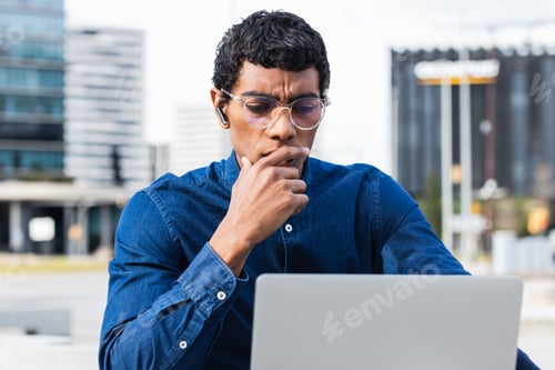 Preview: Handsome man using laptop in a city. Focused indian male student working on computer outdoor.