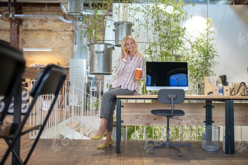 Preview: Blonde woman sitting on desk, talking on phone, holding cup of coffee