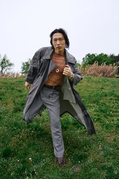 Preview: Stylish young asian man in grey outfit running through a vibrant park on a cloudy day