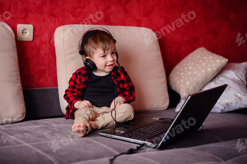 Preview: Little toddler boy sits on sofa wathes cartoons on laptop using headset
