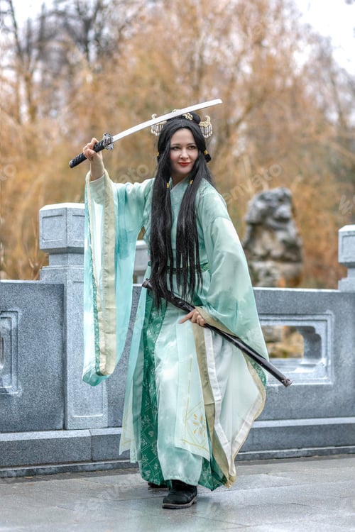 Preview: Asian woman in traditional blue hanfu with katana sword in Chinese park