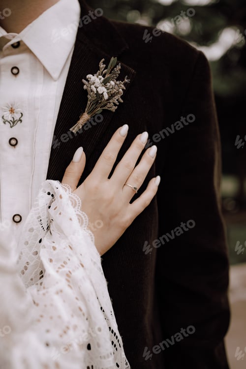Preview: Bride's hand lying on groom's suit with buttonhole.