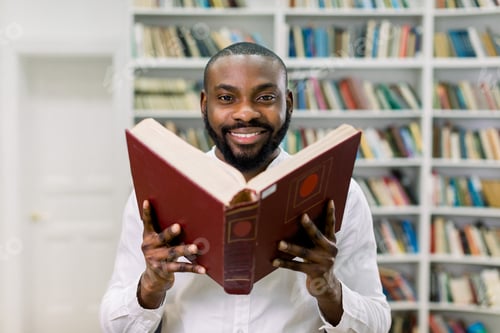 Preview: Good-looking satisfied young African bearded guy in white casual shirt, posing on the book shelves