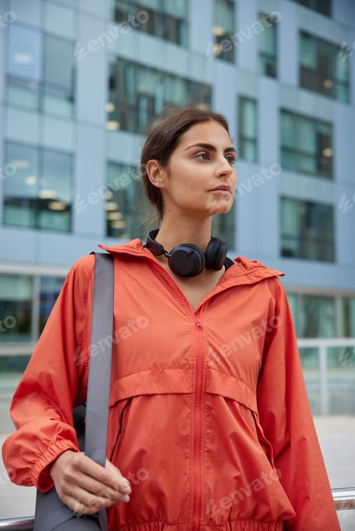 Preview: Vertical shot of brunette woman in red jacket carries fitness mat ready for yoga practice focused in