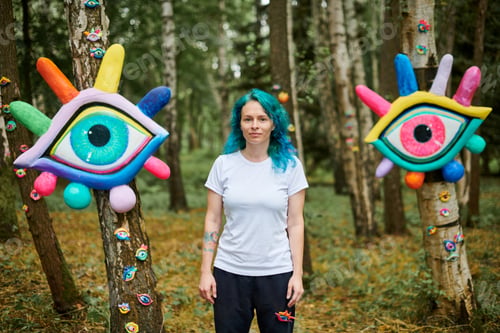 Preview: Weird young adult woman with dyed turquoise hair in white T shirt near big eyes art installation