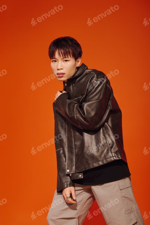 Preview: Stylish young asian man poses confidently against a vibrant orange backdrop in trendy outfit