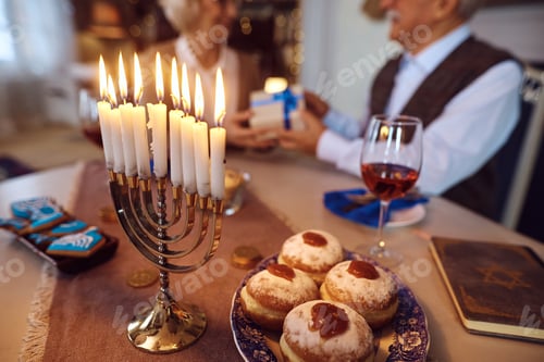 Preview: Close up of menorah with lit candles for Hanukkah with senior Jewish couple in the background.