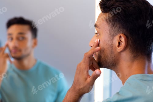 Preview: Biracial man looking at himself in bathroom mirror and applying face cream