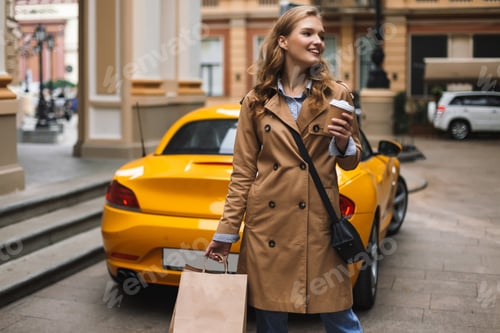 Preview: Young joyful woman in trench coat with little black cross bag ho