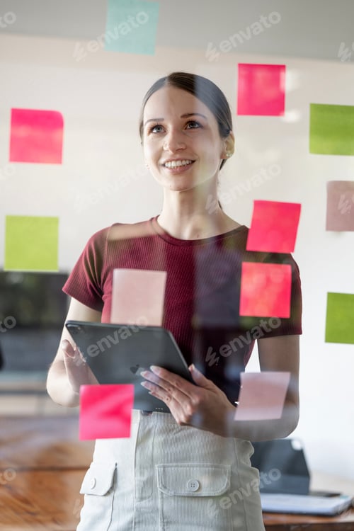 Preview: Young smiling caucasian businesswoman write tasks creative ideas on sticky notes on glass wall.
