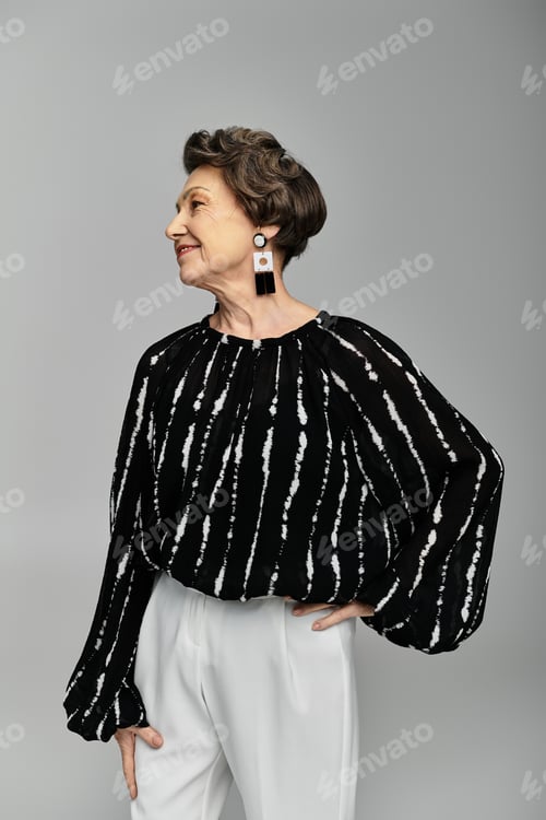 Preview: A beautiful woman in her 60s poses gracefully in trendy attire against a grey backdrop.