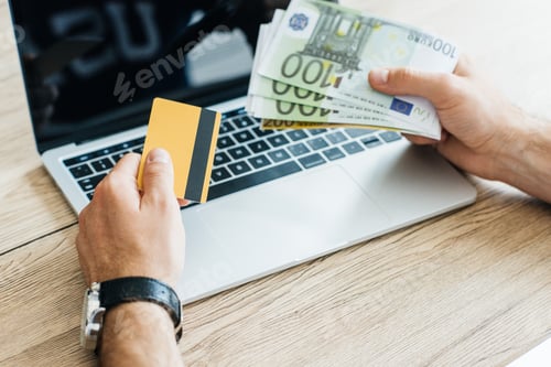 Preview: cropped shot of person holding credit card and euro banknotes above laptop