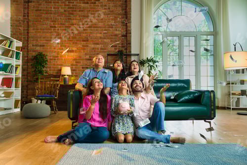 Preview: Indian Asian family of six celebrating a joyful money rain while holding a piggy bank together