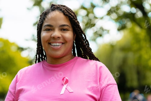 Preview: Young African American woman smiling and wearing pink ribbon for breast cancer awareness, in a park