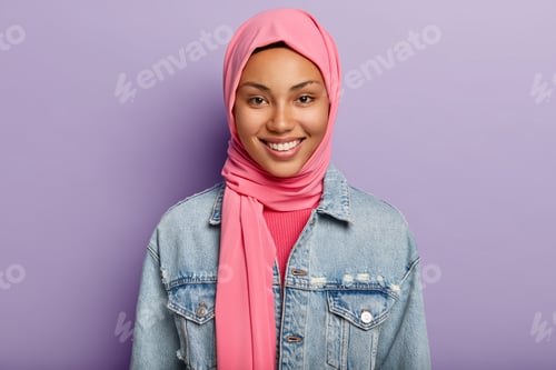 Preview: Studio shot of glad Eastern female has Islamic religion, covered head with pink veil, smiles gently,