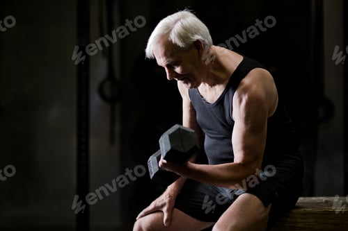 Preview: Senior man sitting doing bicep curls with dumbbell in dark gym