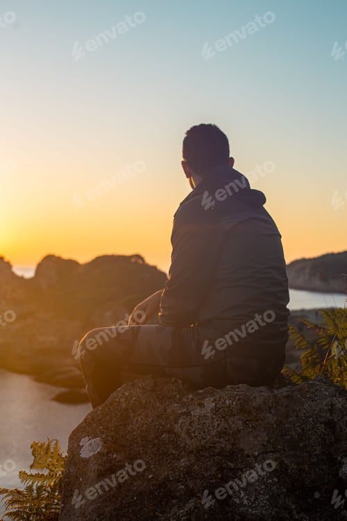 Preview: Vertical shot of a man sitting on a cliff overlooking the ocean and the evening sunset