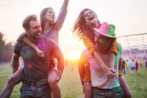 Preview: Two young couples covered in coloured chalk powder piggybacking at Holi Festival