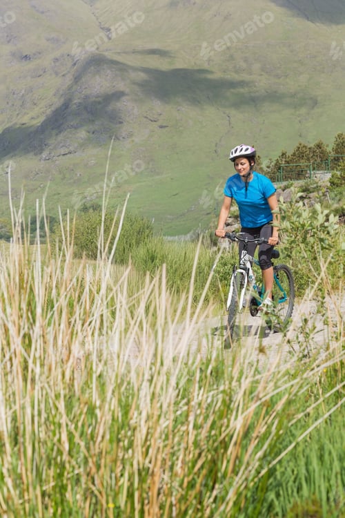Preview: Adult woman riding mountain bike on dirt path through tall grasses in cycling gear, copy space