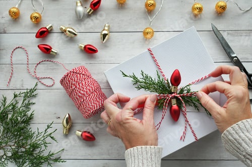 Preview: Woman wrapping Christmas gifts with red & white bakers twine, greenery & red light bulbs, flat lay