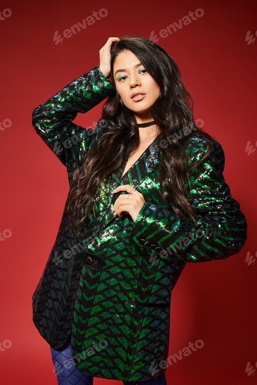 Preview: asian brunette young woman in trendy green jacket with sequins looking at camera on red backdrop