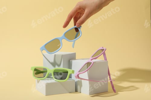Preview: Colorful sunglasses on white blocks and female hand on beige background