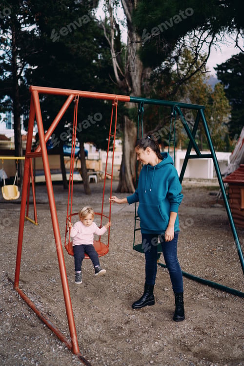 Preview: Mom stands next to the swing on the playground, with a little girl sitting on it